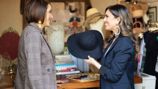 Christine Vartanian standing in a room with racks of clothing and a mirror behind her, wearing a navy blue blazer and large circular pearl earrings, holding a black sun hat, standing in front of a women customer wearing a plaid blazer with gold teardrop earrings