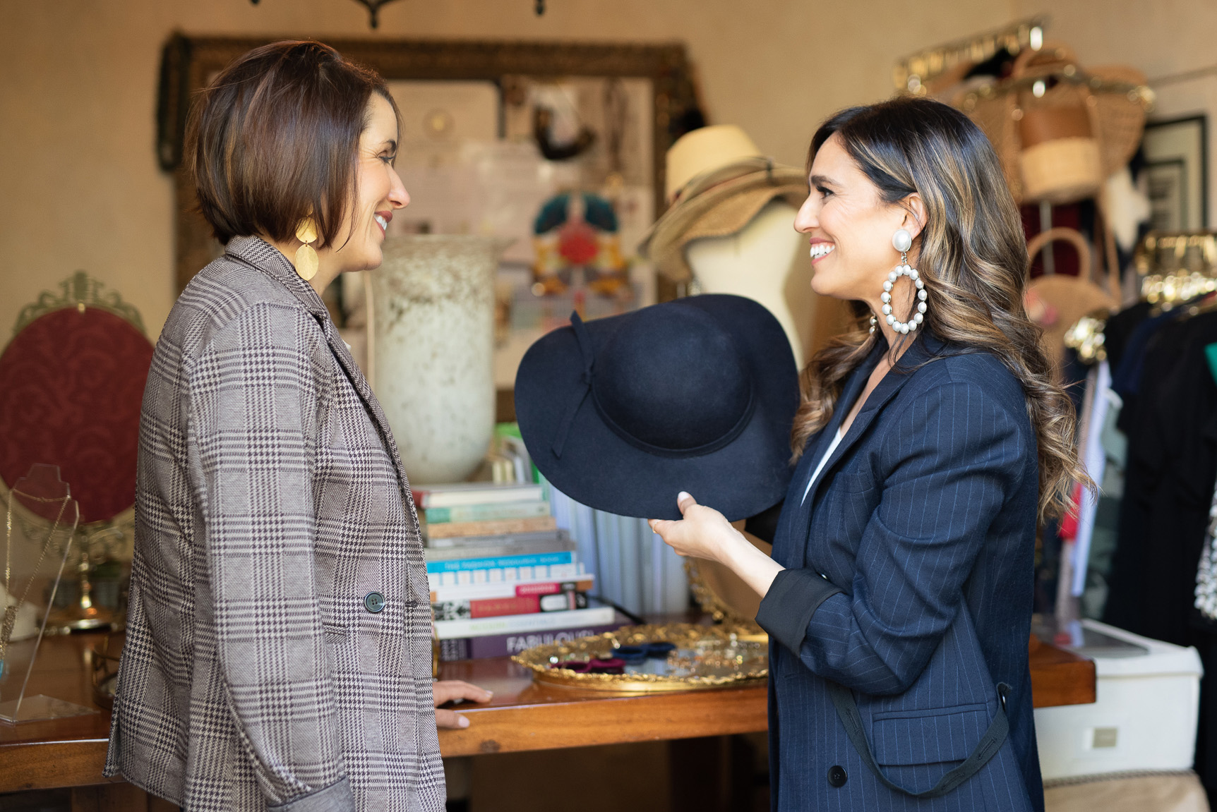 Christine Vartanian standing in a room with racks of clothing and a mirror behind her, wearing a navy blue blazer and large circular pearl earrings, holding a black sun hat, standing in front of a women customer wearing a plaid blazer with gold teardrop earrings