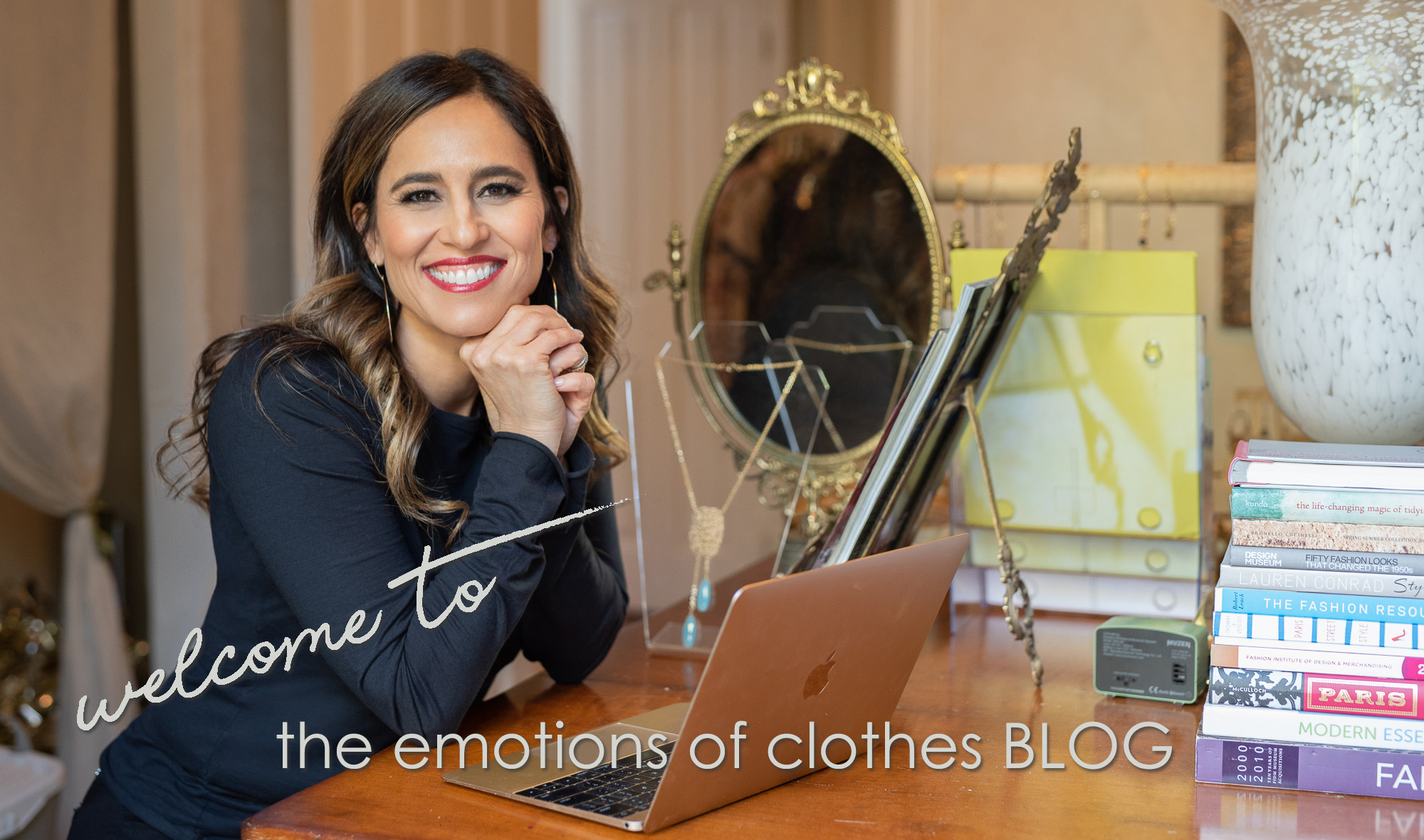 Christine Vartanian wearing a black long sleeved shirt, sitting at a wooden table in front of a laptop with a gold mirror, folders and stack of books on the table, smiling at the camera, with the text "welcome to the emotions of clothes BLOG"