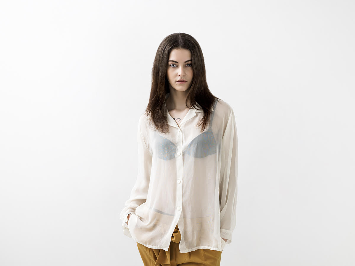 A model with brunette hair standing in front of a white background wearing mustard colored pants, a sheer white top with a black bra underneath staring directly at the camera